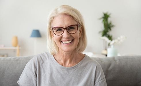 An older woman seated on a couch and smiling after receiving her dental crowns