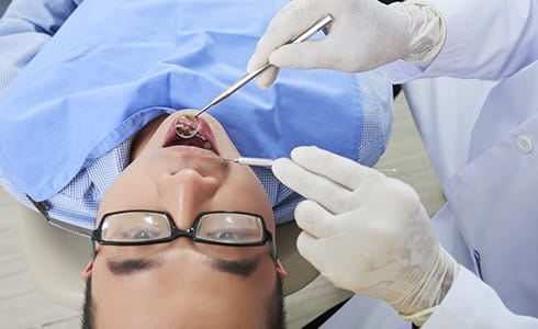 A young male with glasses lying back in a dentist’s chair while a dental professional examines his mouth to determine if he needs a metal-free dental crown