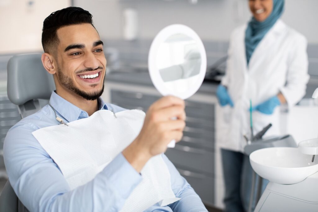 Man in dental chair smiling into mirror with dentist blurry in background